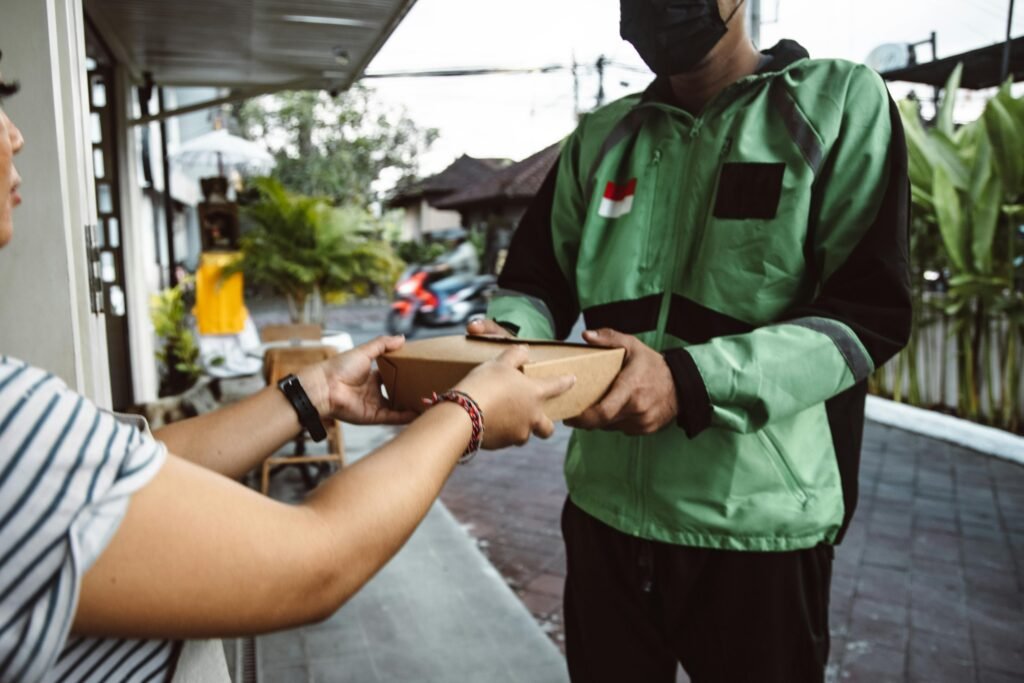 A courier in a green jacket hands a package to a customer outside a house in an urban environment.