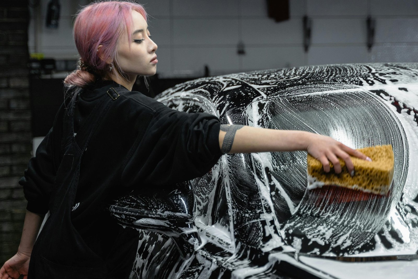A young woman cleans a black car with a sponge and soap in a garage setting, showing care and attention.